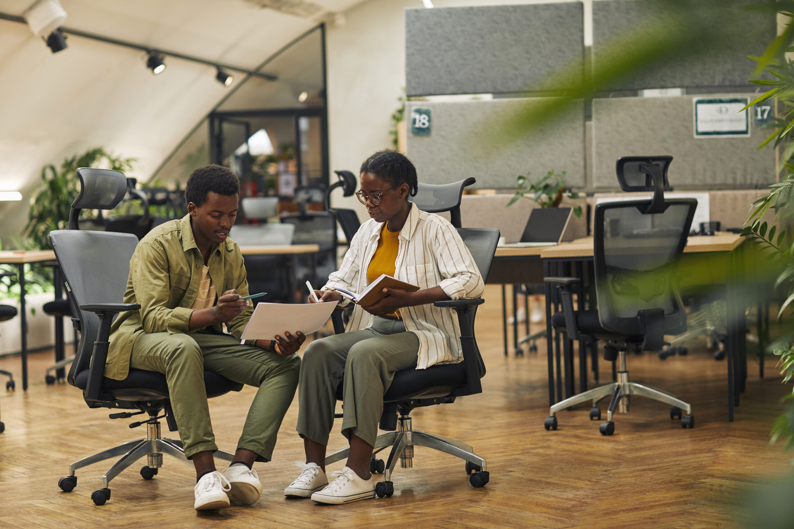 Full length portrait of two contemporary African-American business people discussing work project while sitting on chairs in modern office, copy space