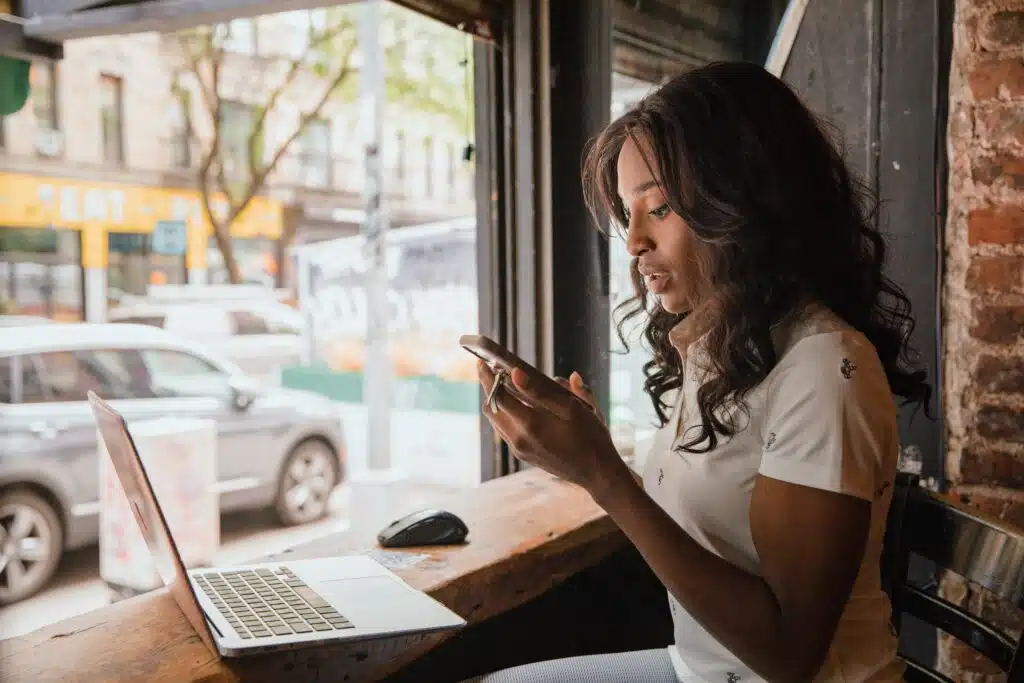 Woman working on a laptop in front of a window while looking at her phone