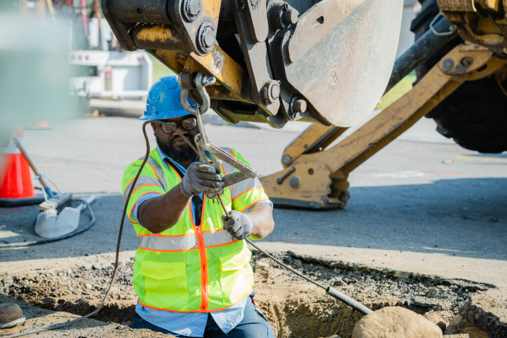 Man wearing hard hat and safety vest using tools to adjust a cable while standing in a hole in the street.