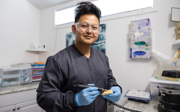 Person wearing medical scrubs and safety goggles holding a dental tool and tooth mold.